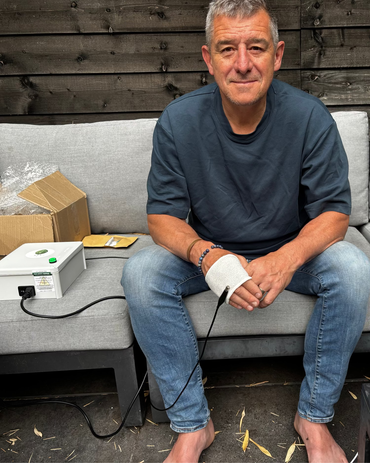 A man sits barefoot on a gray outdoor sofa, wearing a white sleeveless wrist/ankle device connected by a black cable to a small white electronic box on the sofa. A cardboard box and a smartphone rest nearby against a dark wood plank wall.
