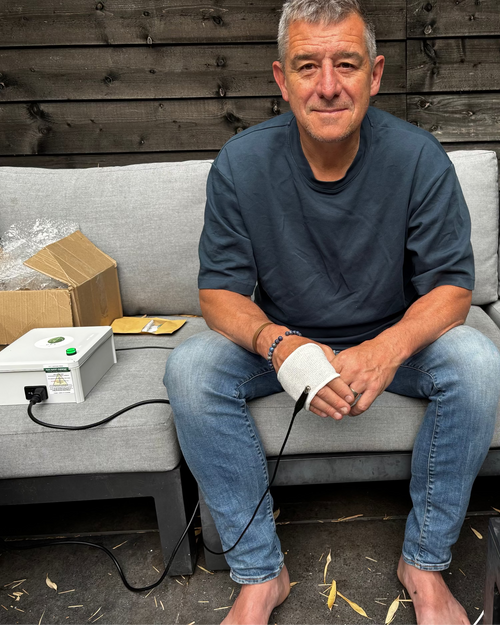 A man sits barefoot on a gray outdoor sofa, wearing a white sleeveless wrist/ankle device connected by a black cable to a small white electronic box on the sofa. A cardboard box and a smartphone rest nearby against a dark wood plank wall.
